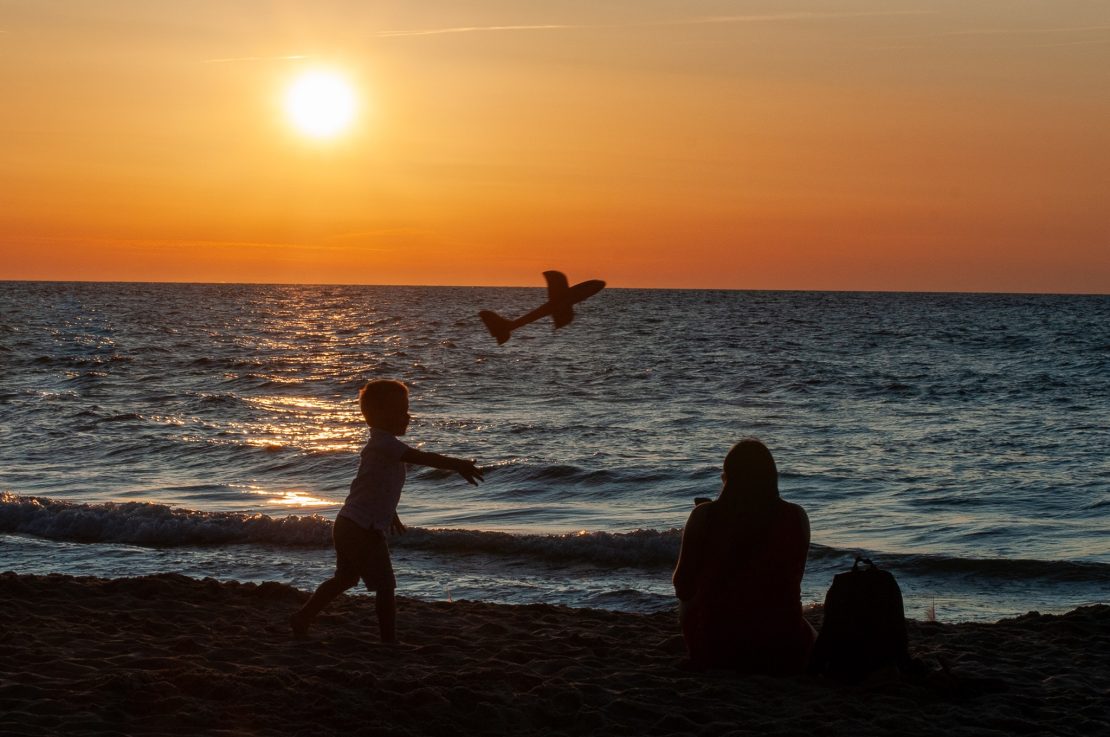 zachód słońca dziecko puszcza samolot obok matka robiąca zdjęcie (ciemne sylwetki na tle zachodzącego słońca )
